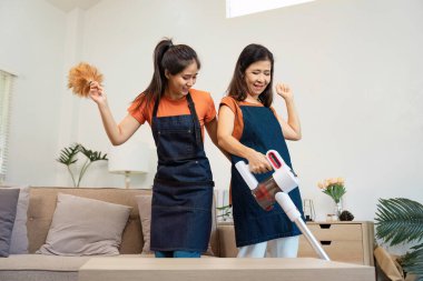 Önlük giyen iki kadın oturma odasını temizliyor, biri elektrikli süpürge kullanırken diğeri toz beziyle neşeyle dans ediyor..
