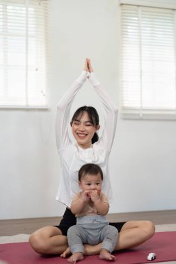 Neşeli bir anne yoga yaparken bebeği neşeyle önde oturur. Huzurlu bir ortamda refahı ve farkındalığı somutlaştırır..