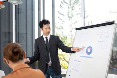 Meslektaşları parlak bir ofiste ortak bir tartışma içindeyken odaklanmış bir adam iş verilerini ters çizelgeye sunar..