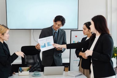 Çağdaş bir ofis ortamında finansal grafikleri işbirliği içinde analiz eden ve stratejileri tartışan çeşitli iş adamları grubu, takım çalışmalarını ve eleştirel düşünceleri vurguladı.