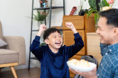 Neşeli bir çocuk heyecanla ellerini kaldırır ve babası ona lezzetli atıştırmalıklar sunar, aile sevgisini ve neşesini gösterir..