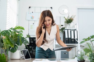 Genç bir iş kadını raporları incelerken gülümsüyor ve modern ofis ortamında telefonla konuşuyor, iletişimi ve takım çalışmasını teşvik ediyor..