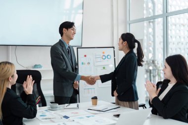 İki meslektaşı, toplantı odasında başarılı bir işbirliği ve takım çalışmasını kutlarken, çok kültürlü bir iş adamı alkışlıyor..
