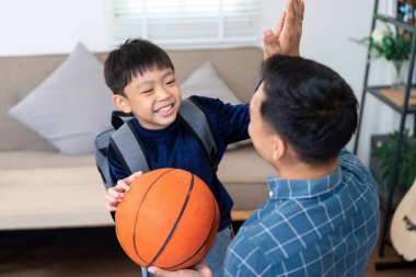 Genç bir çocuk elinde basketbol topuyla okuldan döndükten sonra heyecanla babasını selamlıyor. Onların neşeli etkileşimleri, okula dönüş sezonunda aile desteğinin önemini vurguluyor..