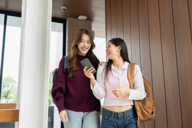 İki arkadaş okul koridorunda yürüyor, kahkahalarını paylaşıyor ve cep telefonlarını kontrol ediyor, öğrenim ve sosyal hayatın karışımını sembolize ediyor..