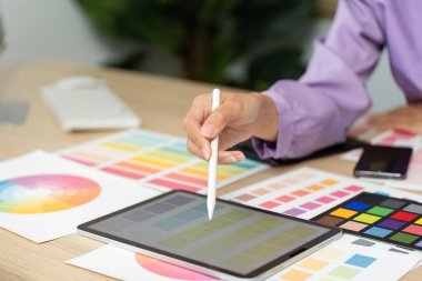 Grafik tasarımcısı, basılı renk paletleri ve tasarım materyalleriyle çevrili, yaratıcılığı ve odak noktasını gösteren bir tablet üzerinde renk seçimi yapar..