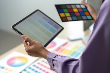 Grafik tasarımcısı, bir tablet üzerindeki dijital renk paletini basılı renk örnekleri ile karşılaştırır, tasarım sürecini ve detaylara dikkati gösterir.