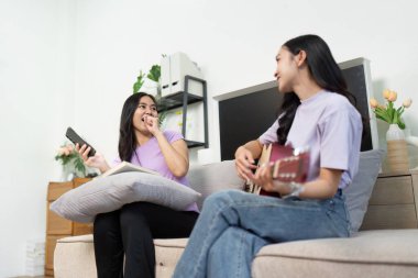 İki genç kadın kanepede keyifli bir anı paylaşırken, biri gitar çalarken diğeri gülümserken, aşk ve arkadaşlık duygusunu yakalıyor..
