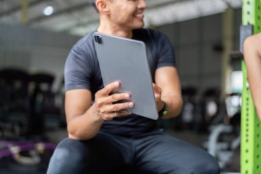 Spor salonunda oturan bir adam, vücut geliştirme ve egzersizlerini takip etmek için tabletle meşgul oluyor..