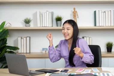 Neşeli bir grafik tasarımcısı, tasarım materyalleri ve dizüstü bilgisayarla çevrelenmiş, coşku ve yaratıcılık içeren başarılı bir projeyi çalışma alanında kutluyor..