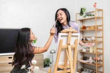 Bir arkadaş diğerinin yüzünü neşe içinde resmediyor, yaratıcılığını ve kahkahalarını canlı bir ortamda gösteriyor..