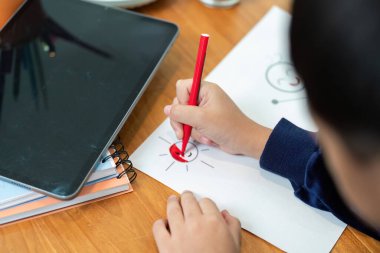 Küçük bir çocuk gülümseyen bir güneşi kağıda çizmek için kırmızı kalem kullanır, bir ev ortamında yaratıcılık ve sanatsal gelişim sergiler..