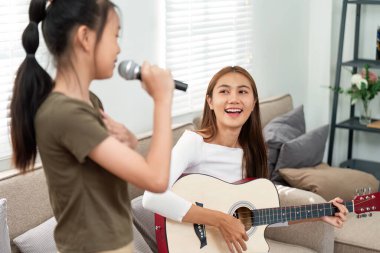Arkadaşı gitar çalarken genç bir kız mikrofona şarkı söylüyor. Oturma odasında neşeli bir müzik ortamı yaratıyor..