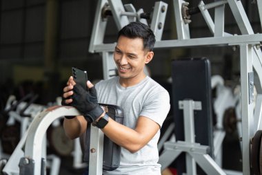 Spor yaparken akıllı telefonuna gülümseyen, spor salonundaki spor salonunun neşesini yakalayan mutlu bir sporcu..