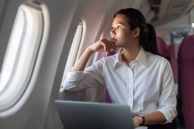 Uçakta dizüstü bilgisayarı üzerinde çalışan odaklanmış bir iş kadını, pencereden dışarı bakarken seyahat sırasında verimlilik sergiliyor..