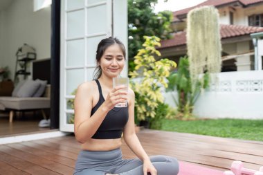 Genç bir kadın elinde bir bardak su tutarken gülümsüyor, yoga seansından sonra serin bir açık hava ortamında hidrasyon ve sağlığını arttırıyor..