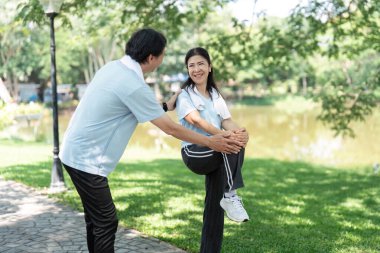 Kıdemli bir adam parkta esneme hareketleri için bir kadına yardım ediyor, yaşlılar için doğal ortamdaki sağlık ve uygunluğun önemini vurguluyor..