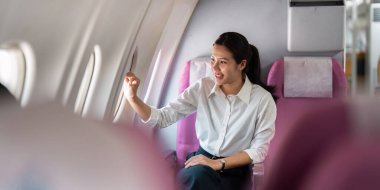 Uçuşundan zevk alan neşeli bir iş kadını uçak penceresiyle etkileşime girerek seyahat deneyimini kucaklıyor..