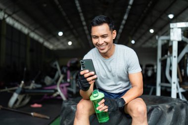 Formda bir adam spor salonunda oturuyor, telefonuna bakıp gülümsüyor. Yoğun bir çalışmadan sonra spor salonundaki başarısını kutluyor..