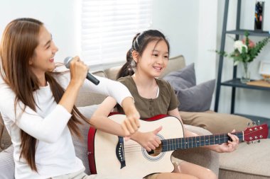 İki kız neşeli bir şarkı söyleme anını paylaşıyor, biri mikrofonlu, diğeri gitar çalıyor, sıcak bir oturma odasında..