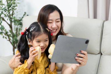 Bir anne ve kızı, tablet üzerinde bir video izlerken, yakın ilişkilerini ve birlikte vakit geçirmenin mutluluğunu vurgulayarak eğlenceli bir anı paylaşıyorlar..