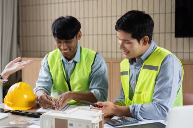 Parlak yelekli iki mühendis yeşil teknoloji projelerinde işbirliği yapıyor, planları gözden geçiriyor ve modern bir çalışma alanında yenilikçi yapı çözümlerini tartışıyorlar..