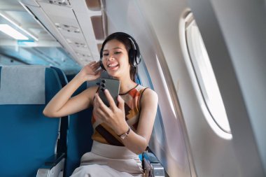 Keyifli bir yolcu bir uçağın penceresinde oturur, kulaklık takarak akıllı telefonunun keyfini çıkarır, modern yolculuğun neşesini somutlaştırır..