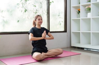 Huzurlu genç bir kadın yoga minderinin üzerinde bağdaş kurarak oturur, elleri dua pozisyonunda meditasyon yapar, farkındalığı ve iç huzuru teşvik eder..