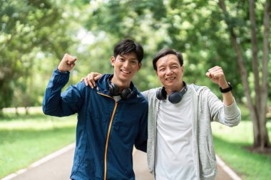 Bir baba ve oğul birlikte poz verir, spor yolculuklarını kutlar ve bereketli bir park ortamında sağlıklı bir yaşam tarzını teşvik ederler..