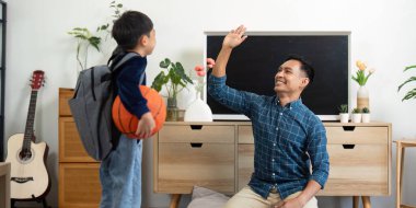 Oğlu el sallarken, bir baba diz çöküp gülümsüyor. Elinde basketbol topu, elinde sırt çantası, aile bağlarını gösteriyor..