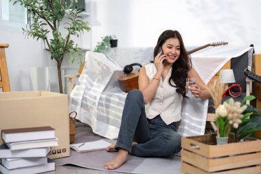 Bir kadın yeni evinin zemininde oturmuş telefonda sohbet ediyor, suyunu yudumluyor ve yeni başlangıcını kucaklıyor..