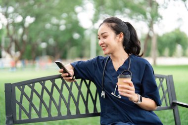 Önlüklü bir doktor bir bankta rahatlar, kahvesini yudumlar ve telefonunu kontrol eder, dengeli bir yaşam tarzını somutlaştırır..