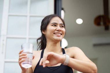Formda bir kadın, elinde bir bardak su tutarken gülümser. Parlak bir iç mekanda egzersiz yaptıktan sonra su ve sağlığını kutlar..