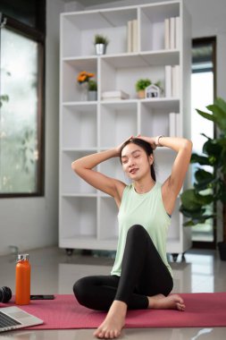 Yoga minderinde oturan genç bir kadın, evinde huzurlu bir atmosferin keyfini çıkarırken esniyor ve dinleniyor..