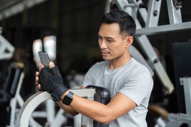 Sporcu ara veriyor, akıllı telefondan spor salonu güncellemelerini kontrol ediyor, teknoloji ile spor rutinlerini modern spor salonunda harmanlıyor..