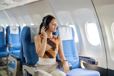 Neşeli bir bayan yolcu, bir uçağın içinde kulaklıkla müzik dinlerken gülümser ve seyahat deneyimini kucaklar..