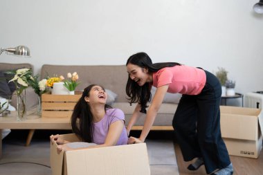 İçeride eğlenen iki neşeli arkadaş, çiçekler ve doğal ışıkla dolu bir oturma odasında karton kutularda gülüp oynuyorlar..