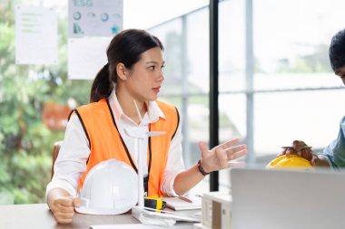 Turuncu yelek giyen bir kadın mühendis, iş yerinde ESG ilkelerini vurgulayarak, bir meslektaşıyla sürdürülebilir mühendislik uygulamalarını tartışıyor..