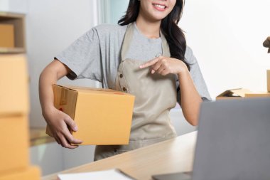 Önlüklü bir girişimci sevkiyatları yönetirken ve paketleri çağdaş bir ofis ortamında teslim etmek için hazırlarken gülümser..