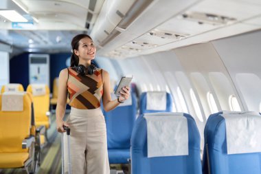 Gülümseyen bir kadın, elinde tabletle bir uçağın koridorunda yürüyor, modern gezgini teknolojiyle somutlaştırıyor..