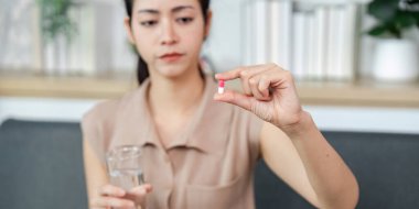 Bir kadın elinde bir bardak su tutarken kapsülü inceler, ilaç seçeneklerini göz önünde bulundurarak..