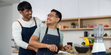 İki adam birlikte yemek hazırlarken keyifli bir anı paylaşıyor. Sıcak bir mutfak ortamında sevgi ve ortaklık sergiliyorlar..