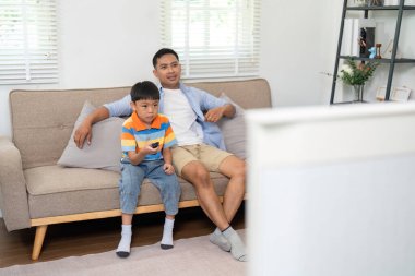 Bir baba ve oğul kanepede birlikte oturur, televizyon izler, rahatlama ve aile bağlarını gösterirler..