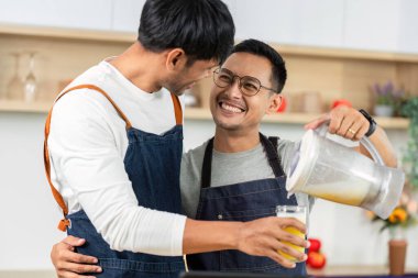 Bir partner gülümserken diğeri meyve suyu dökerken, modern bir mutfak ortamında aşk ve takım çalışmasını ön plana çıkarıyor..