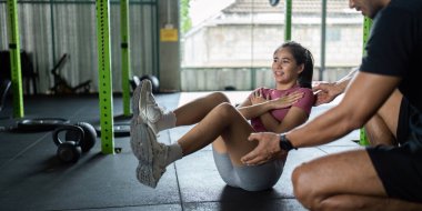 Genç bir kadın spor salonunda antrenörünün rehberliğiyle mekik çekiyor, spor salonunun sağlık durumunu ve desteğini vurguluyor..