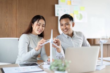 Bir ekip yenilenebilir enerji modellerini tartışıyor, parlak bir ofiste sürdürülebilir çözümler üretirken rüzgar türbini sergiliyor.