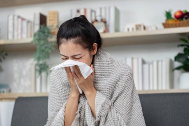 Bir kadın battaniyeye sarılı olarak kanepeye oturup burnunu siliyor ve grip belirtilerini gösteriyor. Sağlığa ve sağlığa odaklan..