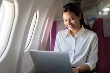 Odaklanmış bir gezgin uçakta otururken dizüstü bilgisayarını kullanır, işi ve seyahati harmanlar. Uzak çalışma ve yaşam tarzı temaları için ideal.