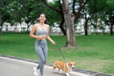 Genç bir kadın yemyeşil bir parkta Corgi 'siyle neşeli bir şekilde koşuyor, aktif yaşam tarzı seçimleriyle fitness ve refahı teşvik ediyor..