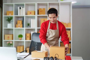 Önlüklü genç bir girişimci, evinde kutu topluyor, e-ticaret işinin operasyonel yönünü gösteriyor..